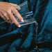 A hand wearing a gold ring pours water onto the Splash Blanket The OG Waterproof Blanket - Small in dark blue micro-velvet, highlighting its elegant look and waterproof comfort.
