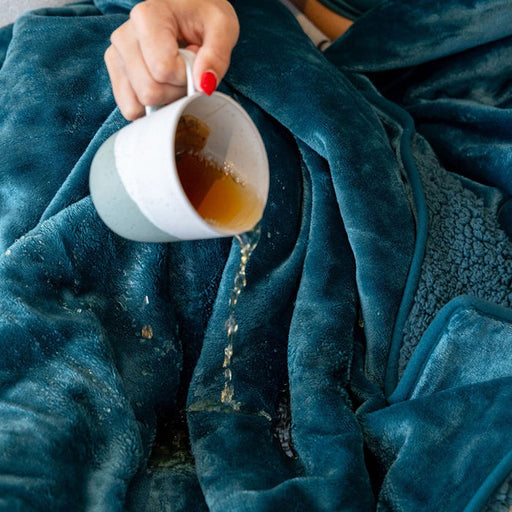 A person with red nail polish spills tea from a white and gray mug onto the Splash Blanket The OG Waterproof Blanket - Small, which features thick, plush micro-velvet in a vibrant teal color.
