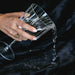 A hand with a gold ring pours water from a clear wine glass onto the Splash Blanket The OG Waterproof Blanket - Medium, showcasing its dark, textured, waterproof surface.