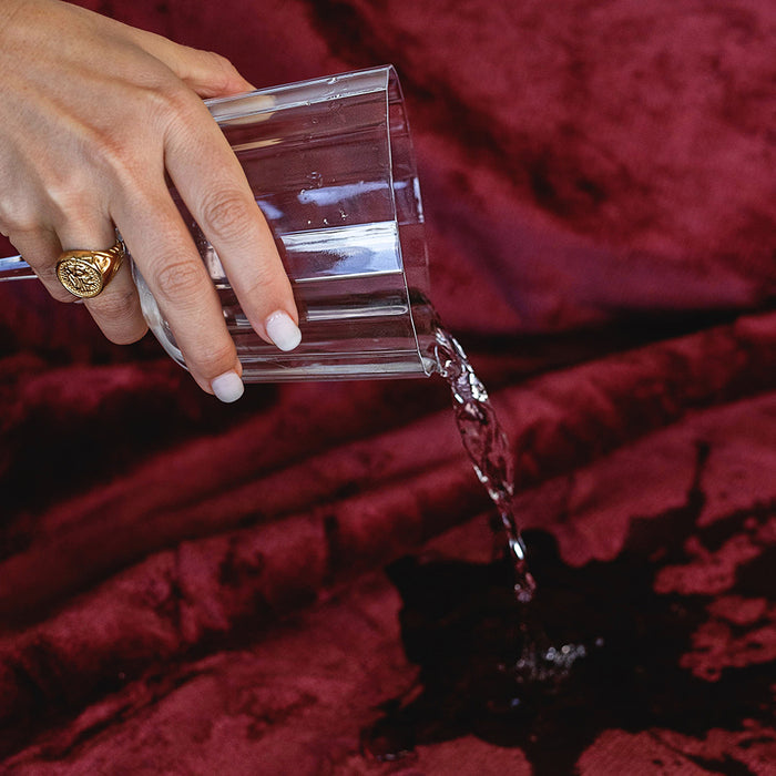 A hand pours water from a glass onto the Splash Blanket The OG Waterproof Blanket - Medium in red velvet, creating a dark spot without soaking through.