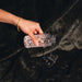 A hand pours water from a patterned glass bottle onto the Splash Blanket Furlush Luxurious Faux Fur Waterproof Blanket - Large, its dark plush surface catching the spill with effortless elegance.