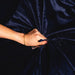 A hand wearing a gold bracelet grips and gathers the Splash Blanket Furlush Luxurious Faux Fur Waterproof Blanket - Large in dark blue velvet, creating elegant wrinkles around the fist.