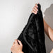 A person displays the Splash Blanket The OG Waterproof Blanket - Large, featuring a soft, plush black surface with neat edging, against a textured white wall.