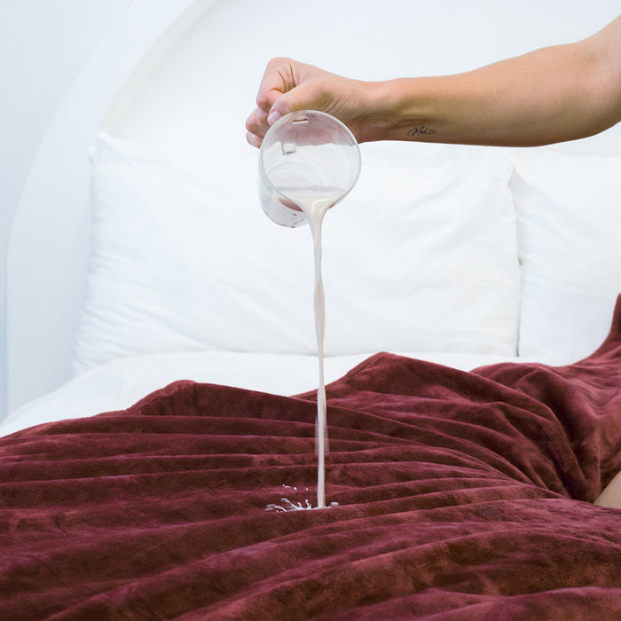 A hand pours white liquid, likely milk, onto the maroon Splash Blanket The OG Waterproof Blanket - Large, spread across a bed with white pillows in the background.