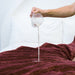 A hand pours white liquid, likely milk, onto the maroon Splash Blanket The OG Waterproof Blanket - Large, spread across a bed with white pillows in the background.