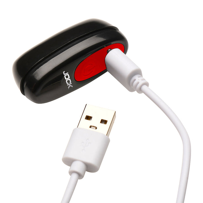 A white USB cable is plugged into the Hard-On Vibrating Silicone Cock Ring with Clit Stim, a small black and red device, against a white background.