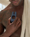 A man holding a Jimmyjane Deimos Dual Motor Vibrating Cock Ring for Couples from Pipedream Products against his chest, featuring a tattoo on his shoulder, partially covered with a sheer white fabric.