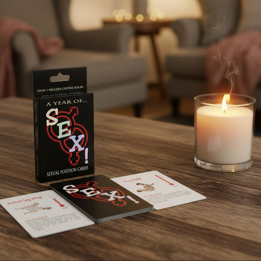 A box of "A Year of Sex! A Sex Positions Card Game" sits on a table in a cozy, warmly lit living room, with three illustrated cards spread out and a lit candle nearby.