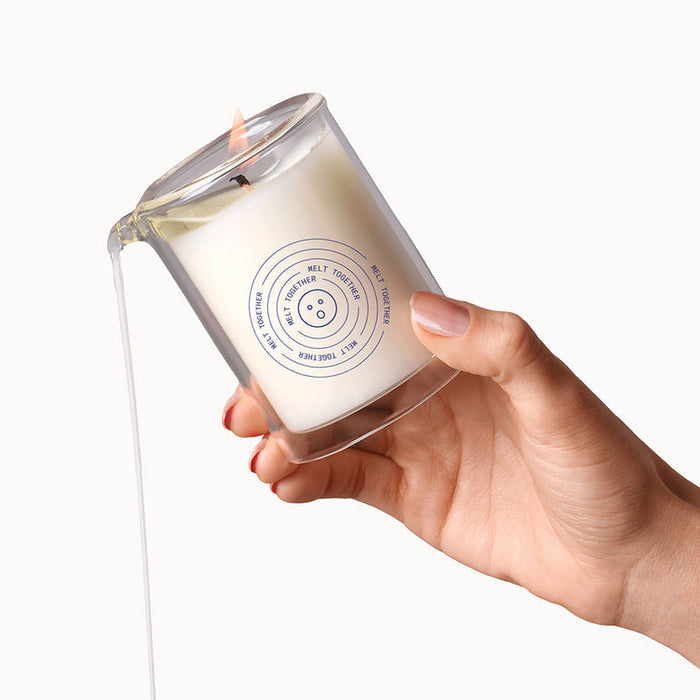 A hand tilts the Soft Touch Massage Oil Candle by Dame (Benzoin, Guaiac Wood & Sandalwood), pouring white, plant-based melted wax from its labeled glass—ideal for moisturizing massages.