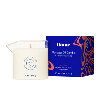 A white, plant-based Soft Touch Massage Oil Candle by Dame (Benzoin, Guaiac Wood & Sandalwood), 5 oz (141 g), sits in a clear glass next to its blue and orange box, designed for intimacy and mood.