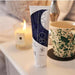 A tube of AH! YES OB Plant-Oil Based Personal Lubricant stands on a table beside a lit candle and a green-speckled mug, with a lamp and part of a pillow visible in the background.