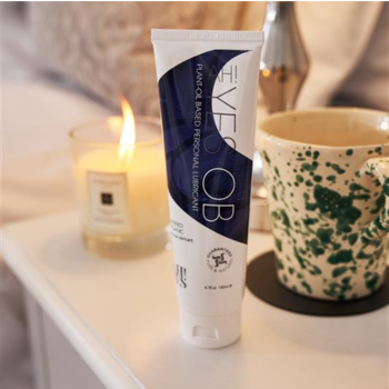 A tube of AH! YES OB Plant-Oil Based Personal Lubricant stands on a table beside a lit candle and a green-speckled mug, with a lamp and part of a pillow visible in the background.