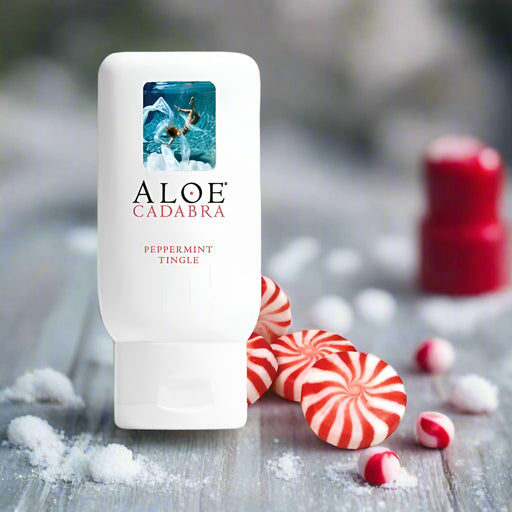 A white bottle of Aloe Cadabra Peppermint Tingle Aloe Based Gentle Lubricant 2.5 oz sits on a surface with peppermint candies and sprinkled sugar, with a blurred red object in the background.