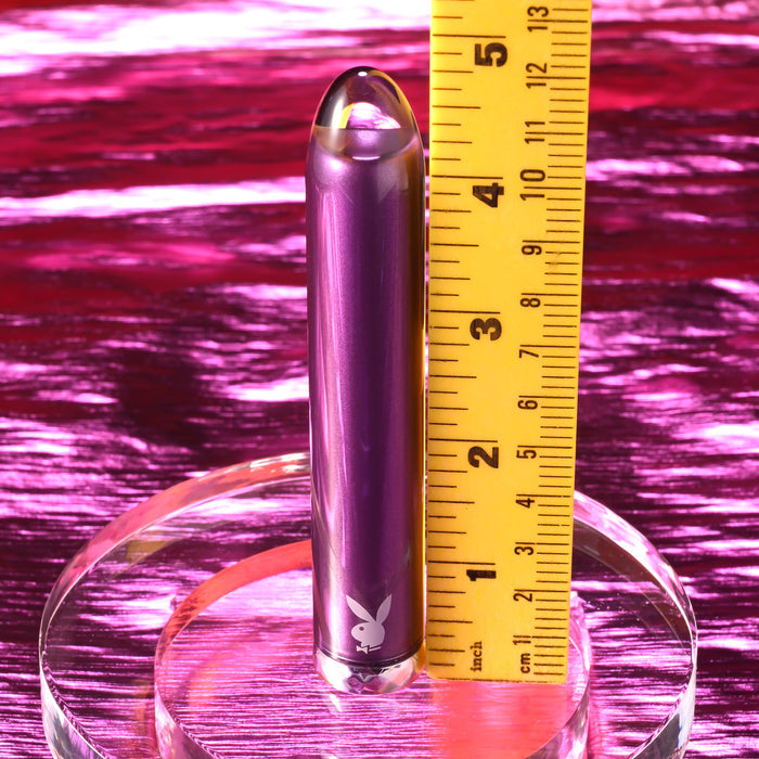 The Amethyst Slim Waterproof Rechargeable Glass Vibrator, featuring a metallic purple finish and the Playboy logo, stands upright on a clear base by a yellow ruler marking its 3.5-inch height, set against a shiny pink background.