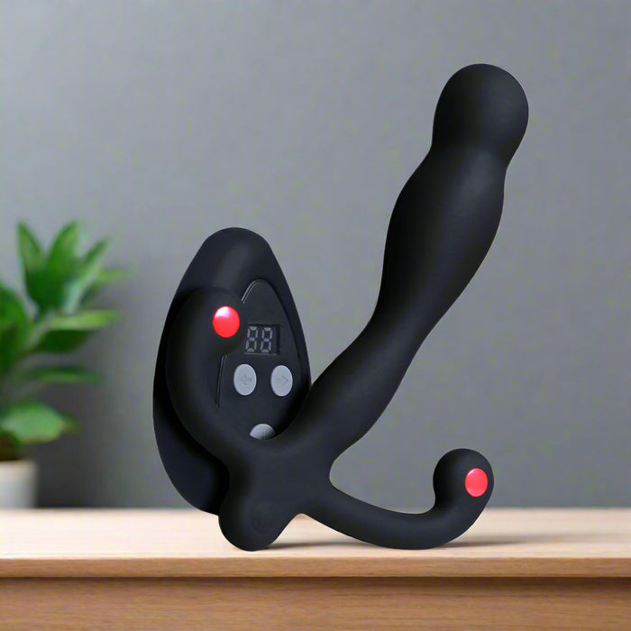 The Aneros Eupho Syn V Vibrating Silicone Prostate Massager with Remote, featuring a curved black silicone design, buttons, and a small display screen, rests on wood. Customizable vibrations and wireless control enhance pleasure. A blurred green potted plant is in the background.