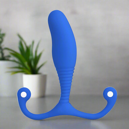 The Aneros MGX Syn Trident Blue Hands-Free Prostate Massager, with its curved ribbed design and two rounded handles, stands upright on a surface. Blurred green potted plants can be seen in the background.