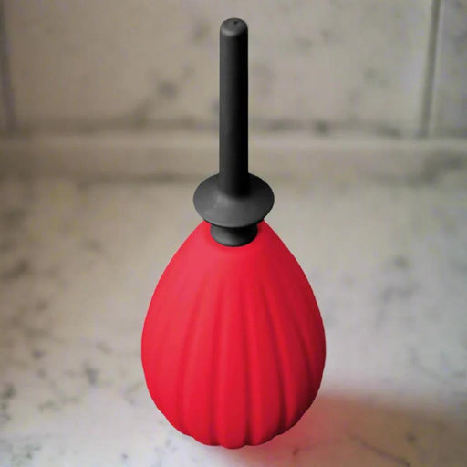 The Aneros Prelude Enema Bulb, featuring a red medical-grade silicone body and black nozzle, stands upright on a white marble surface against a tiled background.