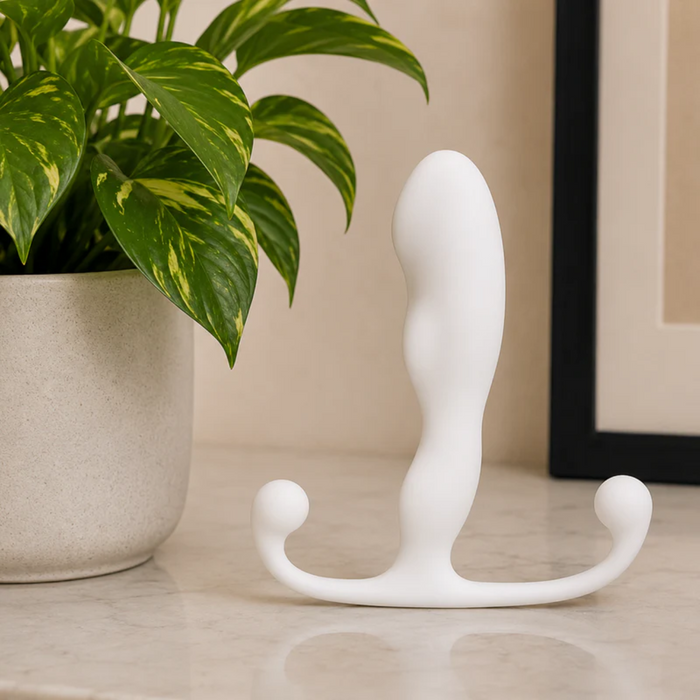 The Aneros Helix Trident Hands-Free Prostate Massager, a white silicone device with two rounded ends and a curved design, sits upright on a marble surface by a potted plant and picture frame, designed for hands-free stimulation.