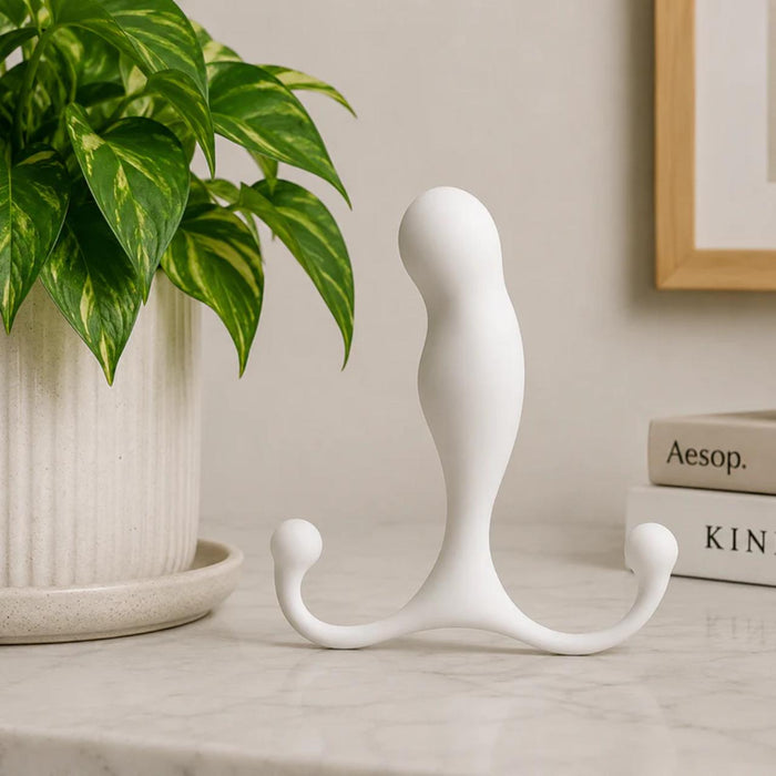A white, curved Aneros MGX Trident Hands-Free Prostate Massager with two rounded arms rests on a marble surface near a green plant and stacked books, suggesting hands-free pleasure and prostate stimulation in a minimalist setting.
