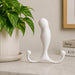 A white, curved Aneros MGX Trident Hands-Free Prostate Massager with two rounded arms rests on a marble surface near a green plant and stacked books, suggesting hands-free pleasure and prostate stimulation in a minimalist setting.