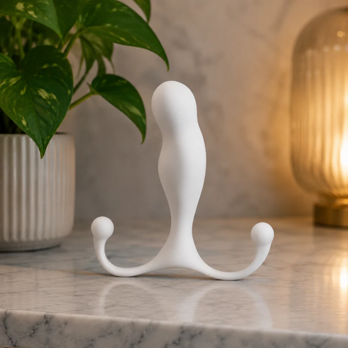 The white Aneros Maximus Trident Hands-Free Prostate Massager, featuring two rounded handles, stands upright on a marble countertop, with a potted plant and glowing lamp in the background for a cozy ambiance.