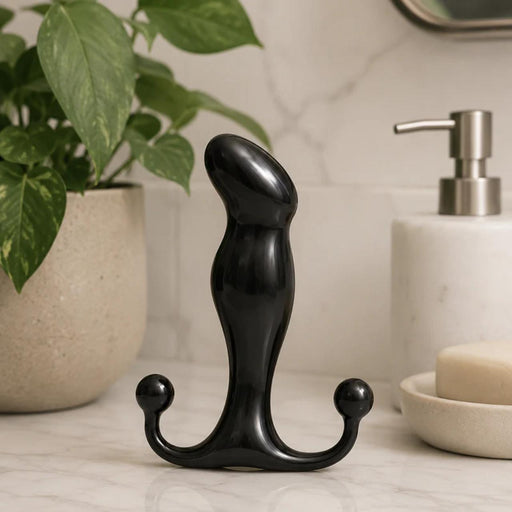 A black Aneros Progasm Jr. Hands-Free Prostate Massager stands upright on a marble bathroom countertop next to a potted plant, soap dispenser, and bar of soap—ideal for hands-free prostate stimulation.