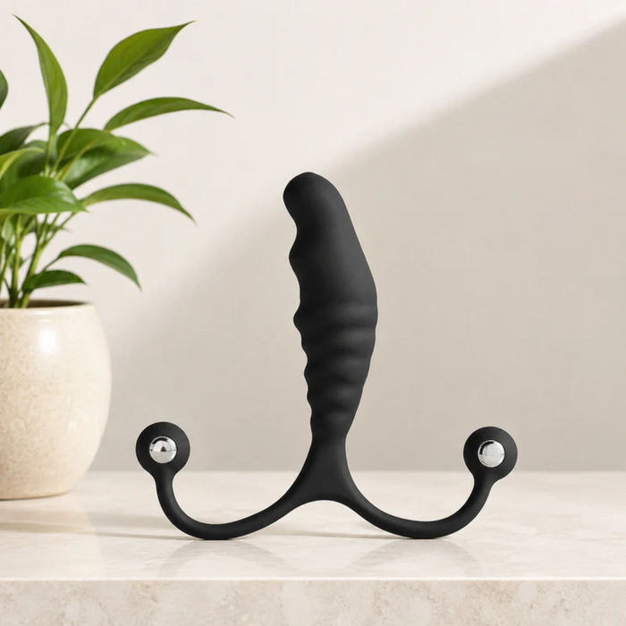 The Aneros Psy Adjustable Prostate Massager, a black textured stimulator with dual round handles, stands upright on a marble surface. A green potted plant is in the background, and bright lighting highlights its hands-free design.