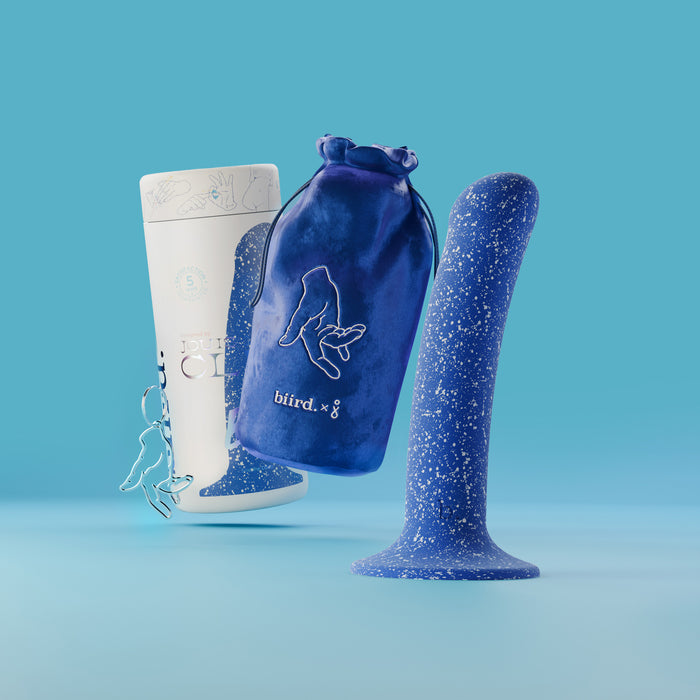 The Bae Slim Smooth 6 Inch Silicone Suction Cup Dildo, in blue speckle, stands upright beside a soft blue pouch with a bird design and a white bottle on a light blue background. This silicone toy is strap-on compatible for versatile pleasure.
