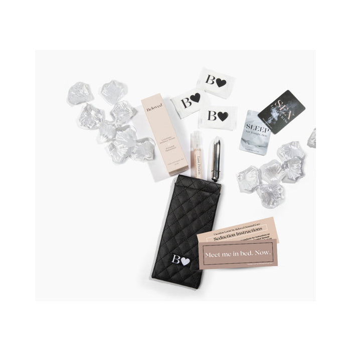 A flat lay displays the Beloved Vacation Lovin’ Intimacy Kit’s black quilted pouch, lip balm, product boxes, business cards, clear acrylic gems, and promotional cards—artfully arranged on a white background.