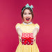 A joyful woman in a yellow dress and green bow headband extends a red polka-dotted gift box with excitement against a pink background, ready to redeem her Giftwizard's gift card.