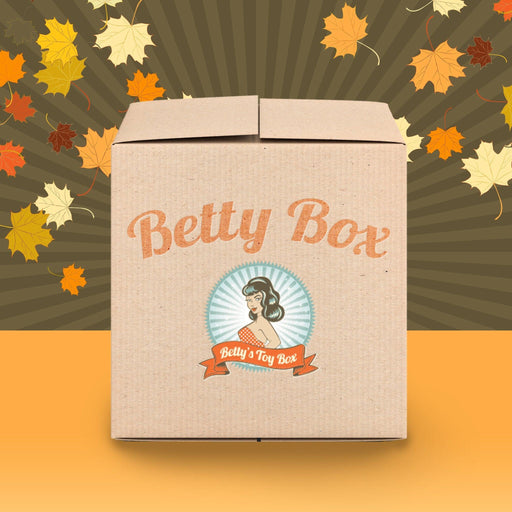 A brown cardboard box with "Betty's Toy Box" written on it in cursive orange letters. Below the text is an illustration of a woman with a vintage hairstyle, encircled by a blue background. Surrounding the box are falling autumn leaves against a radial autumn-themed backdrop, promising the thrill of exploration within: Betty's Boundless Booty Anal Sex Mystery Box - Fall/Winter Edition.