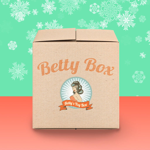 A cardboard box featuring the "Betty Box" logo in a retro-style font on one side. Beneath it, an illustration of a woman is complemented by a ribbon reading "Betty's Toy Box." The background boasts a green top adorned with snowflakes and a red bottom, offering a sneak peek into the exciting contents of the Betty’s Boundless Booty Anal Sex Mystery Box - Holiday 2024 Edition.