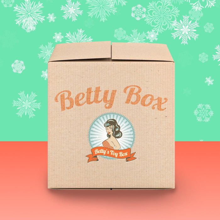 A cardboard box labeled "Betty's Kinky Kitties Mystery Box - Holiday 2025 Edition" features a retro woman, a festive green background with white snowflakes, and rests on a coral-colored surface, hinting at an adult holiday surprise.