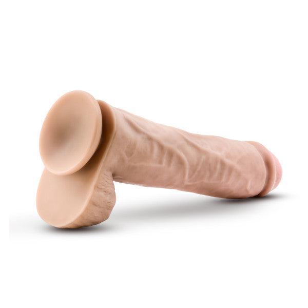 The Big John 11.5 Inch Dual Density Suction Cup Dildo - Vanilla by Blush is a realistic-shaped, beige dildo featuring detailed textures. It stands on a flat surface with its base facing forward and is equipped with a sturdy suction cup for hands-free use.