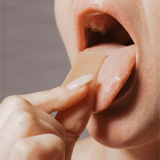 A close-up of a person placing a Bijoux Indiscrets Slow Sex Oral Sex Strip from the 7-Pack onto their tongue, mouth open and tongue out, ready to enjoy a minty cooling sensation.