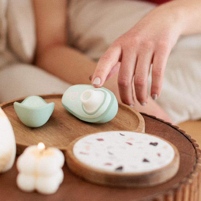 A person's hand reaches for a mint-green Womanizer Liberty 2 Pleasure Air Travel Sized Clitoral Stimulator on a wooden tray, alongside another waterproof vibrator, a decorative coaster, and a lit cloud-shaped candle.
