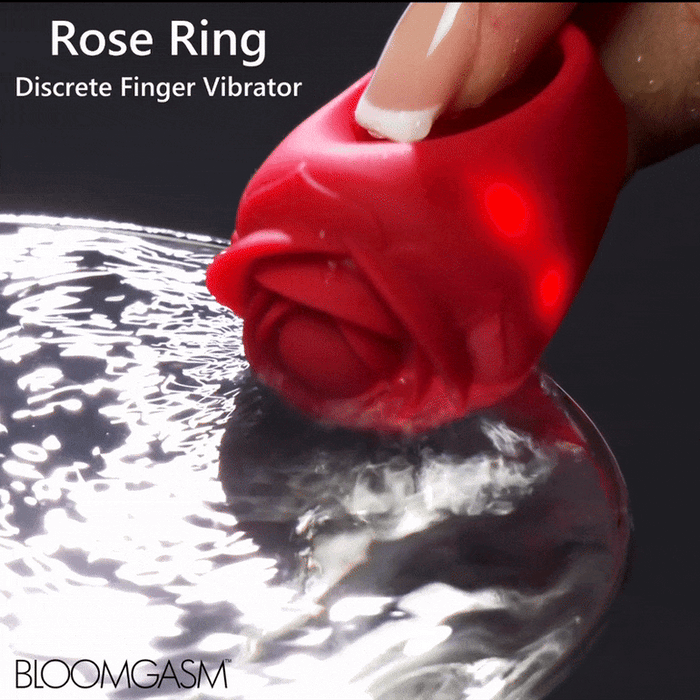 A hand holds a red, rose-shaped wearable vibe made of body-safe silicone above rippling water. The product is labeled "Bloomgasm Rose Ring Finger Vibrator" with the brand "BLOOMGASM" in the lower left corner.
