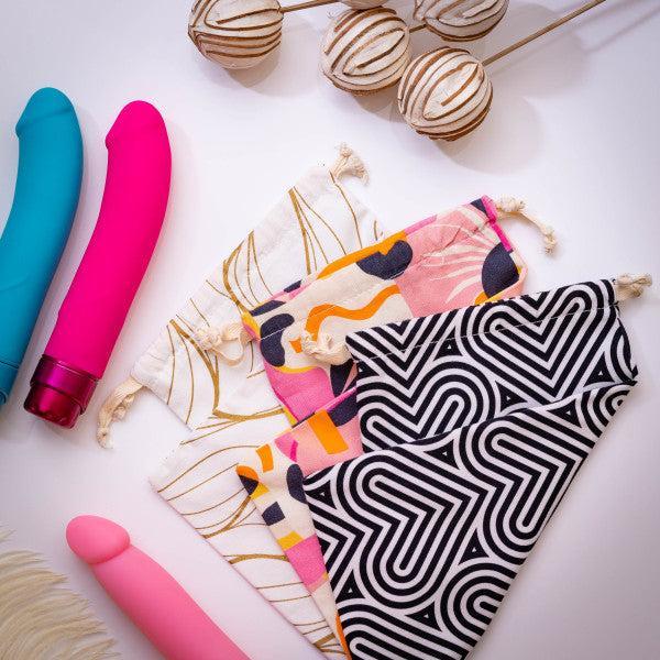 A flat lay photo showcases a variety of vibrantly colored sex toys, including a blue one, a pink one, and a flesh-toned one. Alongside them are three Blush Burst Colorful Toy Storage Bags from the brand Blush, featuring abstract and geometric designs. They are machine washable made from cotton and come with stylish drawstring pouches perfect for sex toy storage. Some decorative dried plants complete the scene.