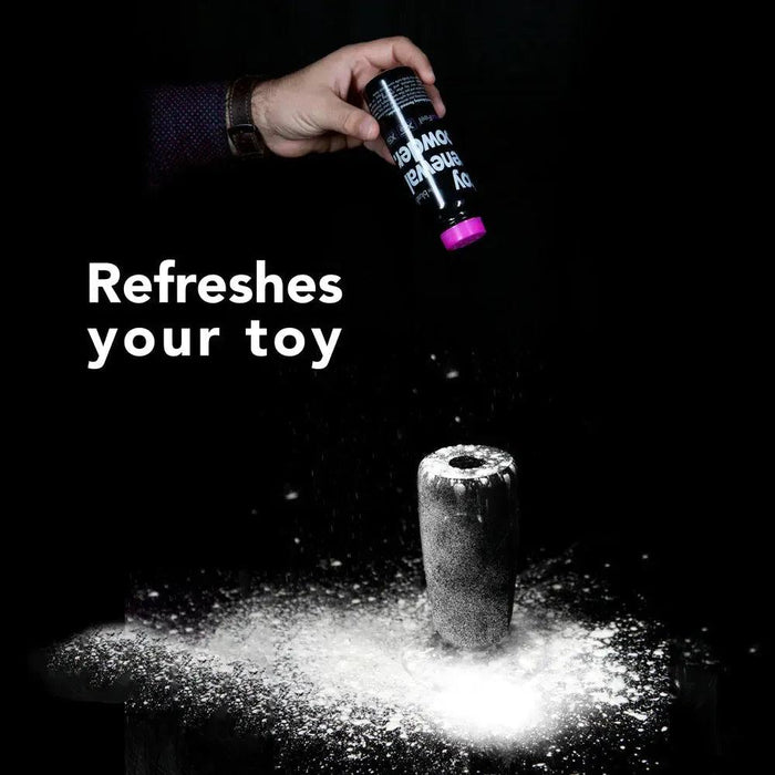 A hand sprinkles Blush SensaFeel & TPE Toy Renewal Powder 3.4 oz from a sleek black bottle labeled in white text onto a cylindrical object against a dark backdrop. Beside the powdery scene, the bold words "Refreshes your toy" are prominently displayed in white.
