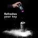 A hand sprinkles Blush SensaFeel & TPE Toy Renewal Powder 3.4 oz from a sleek black bottle labeled in white text onto a cylindrical object against a dark backdrop. Beside the powdery scene, the bold words "Refreshes your toy" are prominently displayed in white.