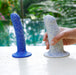 A hand holds the Boo Thick Smooth 5.5 Inch Silicone Suction Cup Dildo in white with blue spots, while both it and a blue version stand upright on a white surface against a backdrop of a blurred pool and green foliage.