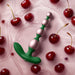 The Bougie Vibrating Aluminum Anal Beads in green and pink rest on a glossy pink surface, surrounded by fresh cherries and cherry juice.
