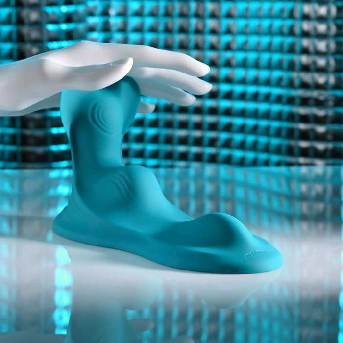 A white mannequin hand rests on the Bumps & Grind Triple Bump Flexible Grinding Vibrator, showcased on a turquoise sensory pad atop a reflective surface, with a blue and silver textured background.