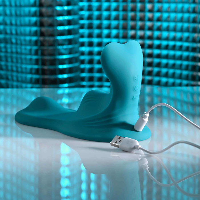 The Bumps & Grind Triple Bump Flexible Grinding Vibrator, a blue liquid silicone adult toy with a curved shape for clitoral stimulation, rests on a shiny surface with its white magnetic USB charging cable nearby against a blue textured background.