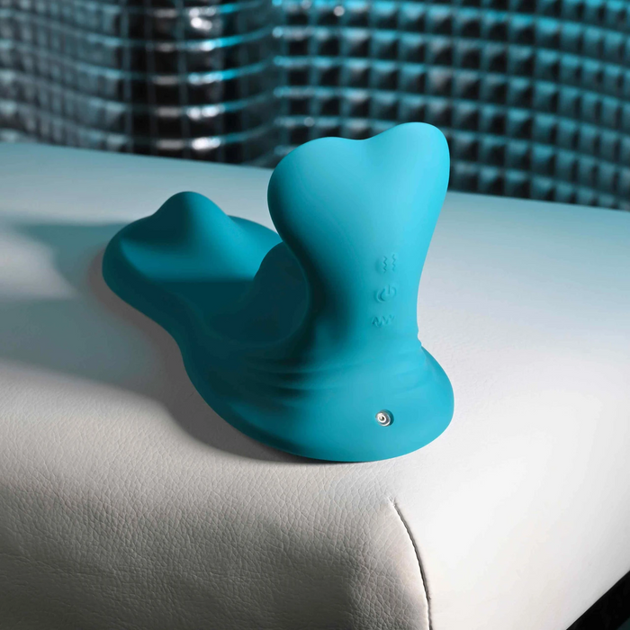 The Bumps & Grind Triple Bump Flexible Grinding Vibrator, a blue silicone personal massager with a curved shape and flexible pleasure pad, sits on white leather with a textured blue background.