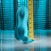 A teal Bumps & Grind Triple Bump Flexible Grinding Vibrator stands upright on a reflective surface next to a yellow measuring tape against a blue textured background, showing the vibrator is about 7 inches tall.