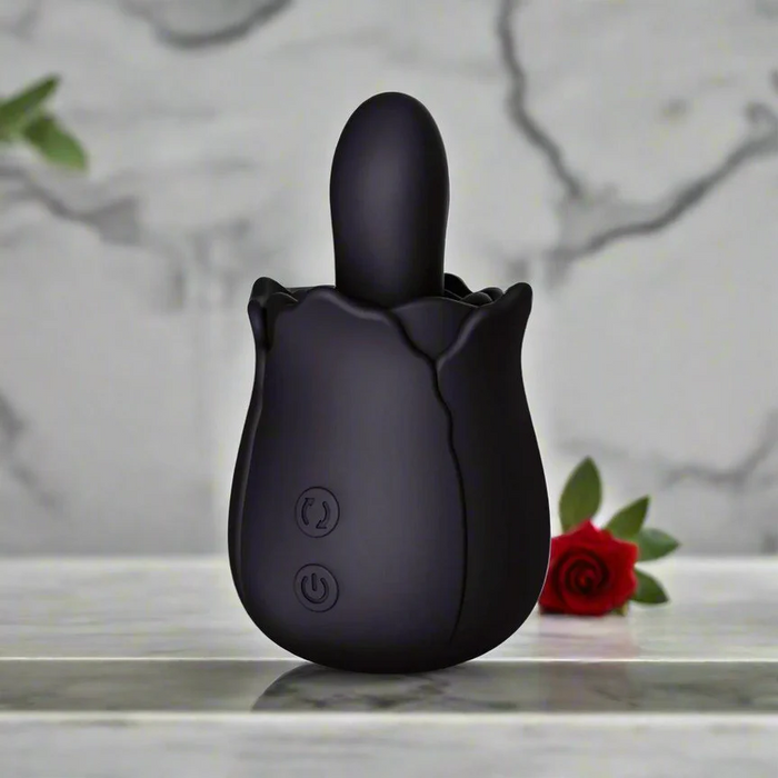 The Bums and Roses Powerful Rimming Tongue Vibrator in black, featuring front control buttons, sits on a marble surface. A blurred red rose and green leaf in the background emphasize its elegant rimming design.