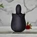 A black, rose-shaped personal massager with two buttons on its base sits on a marble surface. A red rose and green leaves are blurred in the background.