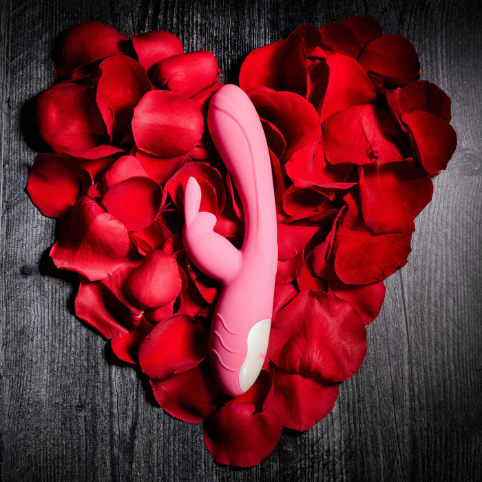 The Bunny Kisses Rechargeable Silicone Rabbit Vibrator in pink rests on a heart-shaped bed of red rose petals, displayed on a dark wooden surface.