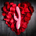 The Bunny Kisses Rechargeable Silicone Rabbit Vibrator in pink rests on a heart-shaped bed of red rose petals, displayed on a dark wooden surface.